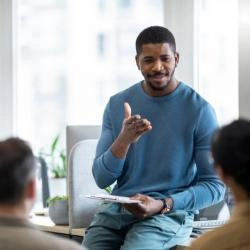 homem-negro-com-uma-prancheta-na-mão-se-comunicando-com-mais-três-pessoas-sentadas